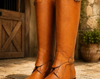May include: A pair of knee-high tan leather boots featuring a smooth finish, a rounded toe, and a low stacked heel. Each boot includes a decorative strap across the ankle and a buckle strap near the top opening, both secured with gold-tone hardware.
