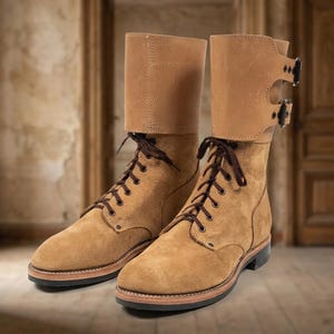 May include: A pair of tan suede boots featuring a high-cut design with dark brown laces and a smooth leather cuff at the top. Each boot includes two adjustable straps with black metal buckles on the outer side and a dark sole with a stacked heel.