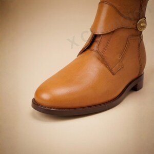 K&ouml;nnte beinhalten: Eine hellbraune Lederstiefelette mit abgerundeter Schuhspitze, einem flachen dunkelbraunen Absatz und einem dekorativen Riemen am oberen Teil, der mit einem runden Metallknopf befestigt ist. Der Stiefel besteht aus glattem Leder mit sichtbaren N&auml;hten.