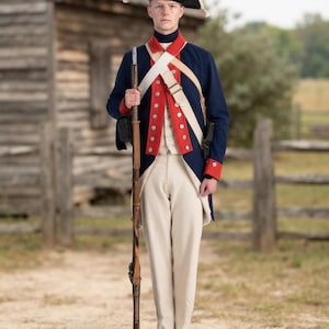 May include: A historical military uniform featuring a dark blue coat with red facings, a white waistcoat, and off-white trousers. The ensemble includes a black tricorne hat, white cross-belts, and a long wooden musket held in a vertical position.