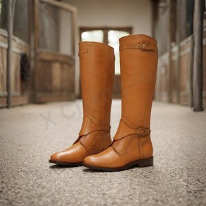 K&ouml;nnte beinhalten: Ein Paar kniehohe Reitstiefel aus hellbraunem Leder mit strukturiertem Schaft, abgerundeter Zehenpartie und einem dekorativen Riemen &uuml;ber dem Kn&ouml;chel, der mit einer kleinen Messingschnalle befestigt ist. Oben befindet sich ein verstellbarer Riemen.