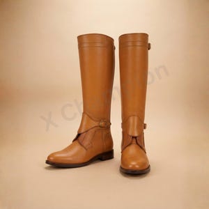 K&ouml;nnte beinhalten: Ein Paar kniehohe Stiefel aus hellbraunem Leder mit glatter Oberfl&auml;che, abgerundeter Zehenpartie und einem dekorativen Riemen mit Metallschnalle im Kn&ouml;chelbereich. Die Stiefel haben einen strukturierten Schaft und einen flachen Absatz.