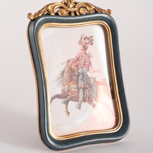 Può includere: Una piccola cornice per foto decorata con una finitura dorata e verde. La cornice contiene un'immagine di una persona a cavallo.