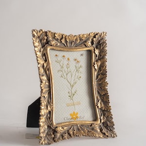 Può includere: Una cornice dorata con un designo ornato a forma di foglia. La cornice contiene un fiore pressato con il testo "Clayre & Eef" in basso.