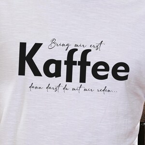Könnte beinhalten: Weißes T-Shirt mit dem deutschen Text "Bring mir erst Kaffee, dann darfst du mit mir reden..." in Schwarz. Das T-Shirt hat einen Rundhalsausschnitt. Die Person trägt eine blaue Jeans.