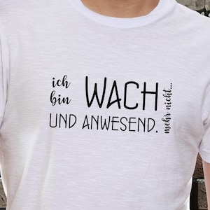 Könnte beinhalten: Weißes T-Shirt mit schwarzem Text: "ich bin WACH und ANWESEND. mehr nicht...". Das T-Shirt wird von einer Person in Jeans getragen. Der Hintergrund ist eine Backsteinmauer.