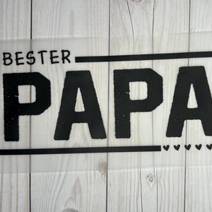 Könnte beinhalten: Ein schwarz-weißes Schild mit dem Text "BESTER PAPA" in fetter Schrift. Das Schild ist mit einem schwarzen Rahmen versehen und hat unten eine Reihe kleiner Herzen.