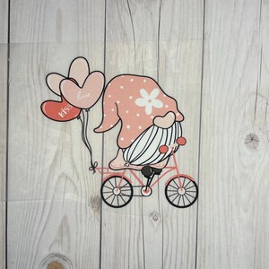 Könnte beinhalten: Ein rosa Zwerg mit weißem Bart und einer rosa-weißen Polka-Dot-Mütze fährt ein rotes Fahrrad mit rosa Herzen, auf denen "love" und "kiss" steht, die am Lenker befestigt sind.