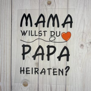 Könnte beinhalten: Ein weißes Schild mit schwarzem Text, auf dem "Mama willst du Papa heiraten?" mit einem roten Herzen steht.