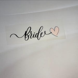 Könnte beinhalten: Ein klarer Aufkleber mit dem Wort "Bride" in eleganter schwarzer Schrift, begleitet von einem hellrosa Herz. Der Aufkleber befindet sich auf einer weißen Oberfläche, mit Blumen und einer Kaffeetasse im Hintergrund.