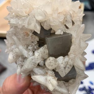 May include: A cluster of clear, crystalline formations with a spiky appearance, surrounding several dark gray, cube-shaped structures. The overall color palette is neutral, with shades of white, gray, and clear.
