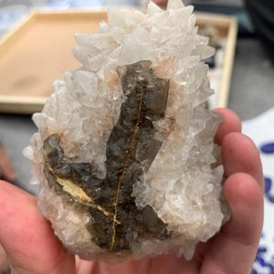 May include: A cluster of natural crystals, primarily white with dark brown and yellow inclusions. The crystals have a rough, textured surface and are held in a hand. The overall shape is irregular and organic, showcasing the raw beauty of the mineral formation.