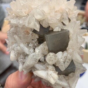 May include: A cluster of clear quartz crystals with sharp, pointed formations. The quartz surrounds several dark gray, cube-shaped pyrite crystals. The overall structure is held in a hand, showcasing the natural beauty of the mineral specimen.
