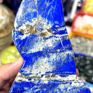 May include: A polished, irregularly shaped lapis lazuli stone. The stone is a deep blue color with white and gold veining throughout. The stone is held in a hand, and other similar stones are visible in the background. This is a decorative item.