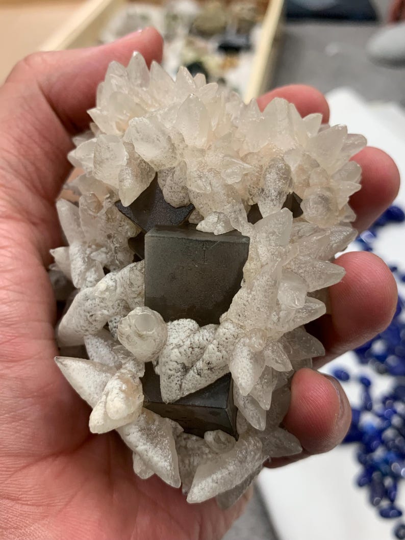 May include: A cluster of clear, crystalline formations surrounding a dark, geometric mineral. The crystals have sharp, pointed edges and a frosted appearance. The mineral appears to be a dark gray or black color. The overall composition is a natural, geological specimen.