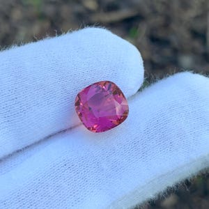 May include: A cushion-cut gemstone with a vibrant pink hue, held against a white fabric background. The gem's facets catch the light, showcasing its clarity and color. The gemstone appears to be a tourmaline or similar precious stone.