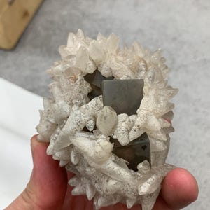 May include: A cluster of quartz crystals with a central, dark, square-shaped mineral formation. The quartz crystals are clear with a frosted appearance, and the overall structure is held in a hand.