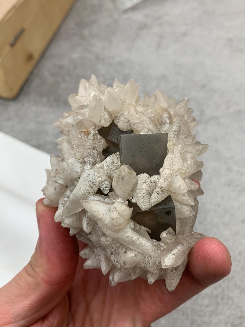 May include: A cluster of quartz crystals with a central, dark, square-shaped mineral formation. The quartz crystals are clear with a frosted appearance, and the overall structure is held in a hand.