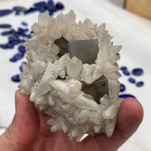 May include: A cluster of quartz crystals with a central, gray, cubic mineral formation. The quartz crystals are clear and pointed, surrounding the gray cubes. The background shows scattered blue gemstones.