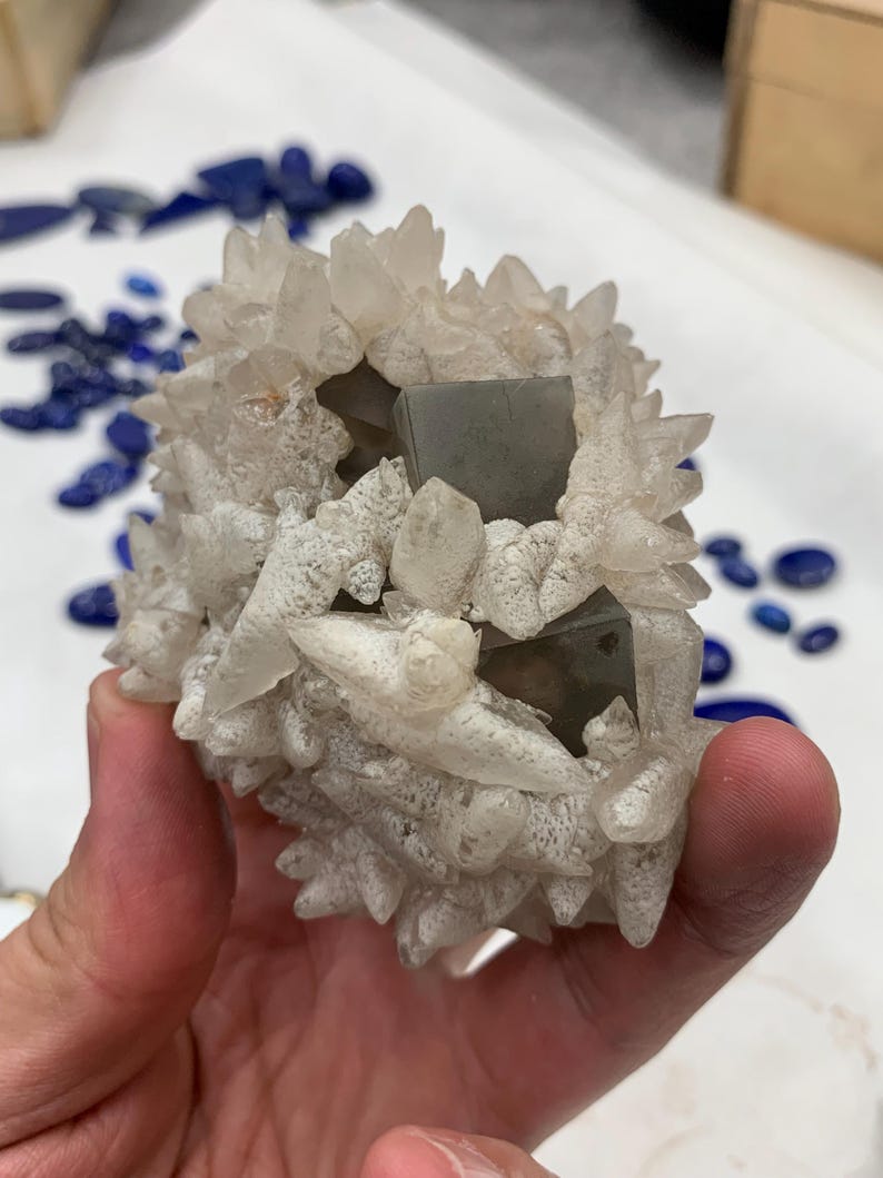 May include: A cluster of quartz crystals with a central, gray, cubic mineral formation. The quartz crystals are clear and pointed, surrounding the gray cubes. The background shows scattered blue gemstones.