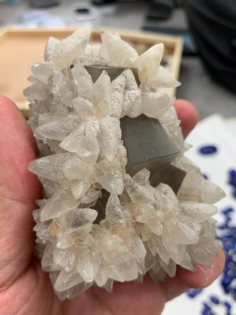 May include: A cluster of clear, crystalline formations with a rough, textured appearance. The crystals are clustered around several dark, square-shaped formations. The overall color palette is neutral, with shades of white, gray, and clear.