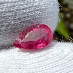 Puede incluir: Una piedra preciosa rosa en forma de pera con facetas. La piedra preciosa está sobre un fondo de tela blanca.