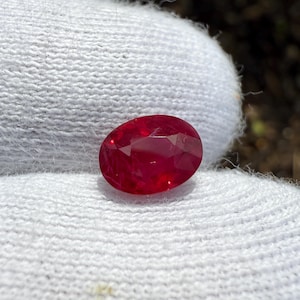 Può includere: Una gemma ovale di colore rosso vibrante con taglio sfaccettato. La gemma è posizionata su uno sfondo bianco.