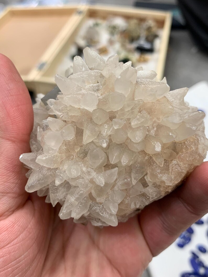 May include: A cluster of clear, crystalline quartz points, held in a hand. The quartz crystals are clustered together, creating a spiky, textured appearance. The base of the cluster has a light tan color. The background shows a wooden box with other crystals.