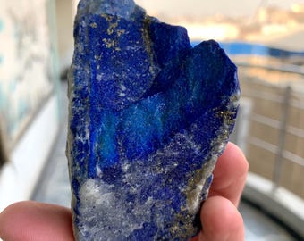 May include: A rough-cut piece of lapis lazuli, a deep blue metamorphic rock with flecks of gold-colored pyrite. The stone is held in a hand, showcasing its natural, irregular shape and rich color variations.