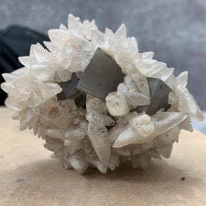 May include: A cluster of clear, crystalline formations with a central, dark gray, cubic structure. The crystals have a pointed, spiky appearance, creating a textured, natural aesthetic. The mineral specimen is set against a neutral background.