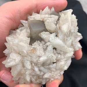 May include: A cluster of light gray and clear crystals, held in a hand. The crystals have a spiky, textured appearance, with a central, geometric, gray crystal formation. The overall shape is rounded and irregular.