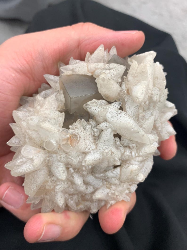 May include: A cluster of light gray and clear crystals, held in a hand. The crystals have a spiky, textured appearance, with a central, geometric, gray crystal formation. The overall shape is rounded and irregular.