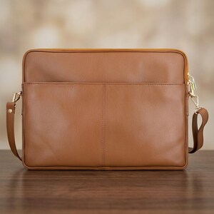 Op de afbeelding: Een rechthoekige, bruinleren laptoptas met een voorvak en een gouden rits. De tas heeft een afneembare schouderriem en goudkleurige hardware. De tas staat op een houten oppervlak.