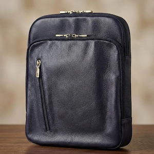May include: A navy blue leather crossbody bag with gold-toned zips. The bag is rectangular with a front pocket and a top zip closure. It is on a wooden surface.