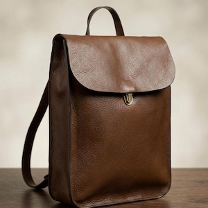 May include: A brown leather backpack with a flap closure and a top handle. The backpack has adjustable straps and a brass-colored clasp. The leather has a textured appearance.