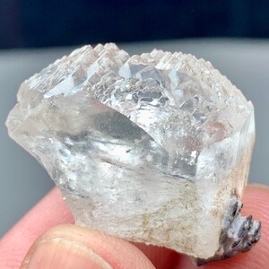 May include: A clear quartz crystal cluster with a rough, textured surface. The crystal has a natural, irregular shape and a light, translucent appearance. The base of the crystal has a dark, speckled texture.