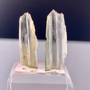 Pode incluir: Duas formações de cristal verde claro, possivelmente turmalina, são exibidas em um suporte de acrílico transparente. Os cristais têm uma estrutura vertical e colunar com aparência translúcida. O suporte é segurado por uma mão.