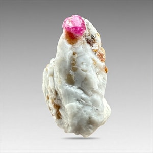 May include: A rough, white mineral specimen with a vibrant pink crystal formation at the top. The crystal has a faceted appearance, and the white base rock has areas of tan and brown coloration.