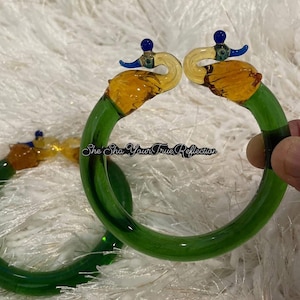Puede incluir: Brazaletes de vidrio verde con detalles en forma de cisne en vidrio amarillo, azul y transparente. Los brazaletes tienen un acabado liso y brillante y se exhiben sobre una superficie blanca y esponjosa. El texto "She Sha Your True Reflection" es visible.