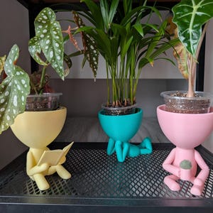 May include: Three potted plants in unique, colourful, human-shaped planters. The planters are sitting on a black metal shelf. The planter on the left is yellow and has a book in its hands. The planter in the middle is turquoise and has a pair of legs. The planter on the right is pink and has a small plant in its hands.