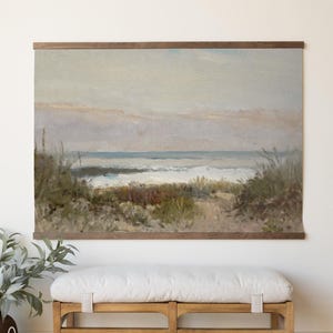 May include: A framed landscape painting of a beach scene with a wooden frame. The artwork depicts a sandy path leading to the ocean, with waves crashing on the shore. The painting is mounted above a wooden bench with a white cushion.