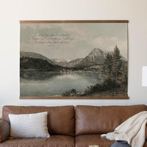 May include: A large landscape wall hanging with a mountain and lake scene, displayed above a brown leather sofa. The artwork has a wooden frame and features a quote in script. The room includes a side table, lamp, and a rug.