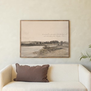 May include: A living room scene featuring a cream-colored sofa with a brown patterned pillow. A framed landscape art piece hangs above the sofa. A round ottoman with books and a small side table with candles complete the decor.