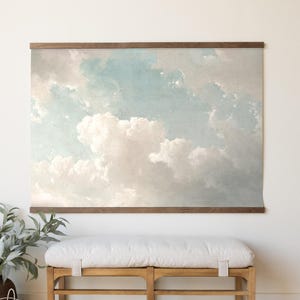May include: A large wall art print featuring a serene sky with fluffy white clouds against a light blue backdrop. The artwork is framed by a dark wooden border and hangs above a wooden bench with a white cushion. A potted plant is on the left.