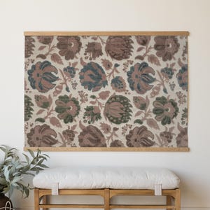 May include: Floral fabric wall hanging with a wooden frame. The fabric features a repeating pattern of large flowers in shades of brown, green, and blue on a cream background. The wooden frame is a light brown color. A wooden bench with a white cushion is below.