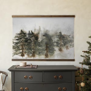 May include: A watercolor print of a forest scene, featuring evergreen trees in shades of green and brown, mounted on a wooden frame. The artwork hangs above a dark gray dresser with ornate bronze knobs. A Christmas tree with ornaments is in the background.
