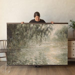 May include: A large, framed art print featuring a landscape painting with a river and weeping trees. The artwork is dominated by shades of green, gray, and white, with a dark brown frame. The print is held up by a person.