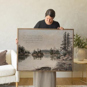 May include: A framed landscape print featuring a serene lake scene with trees and islands. The artwork has a muted color palette and a wooden frame. Text is visible in the upper left corner. A woman is holding the framed print.