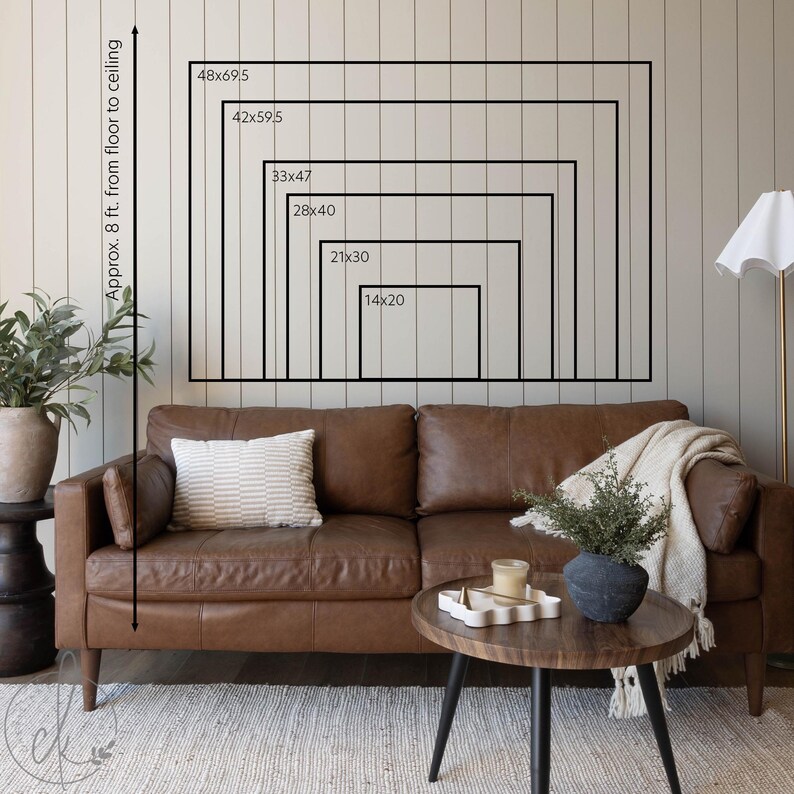 May include: A brown leather sofa with a striped pillow and a cream-colored throw blanket. A wooden coffee table with a decorative tray and a black vase with greenery. The wall displays framed rectangles with dimensions from 14x20 inches to 48x69.5 inches.