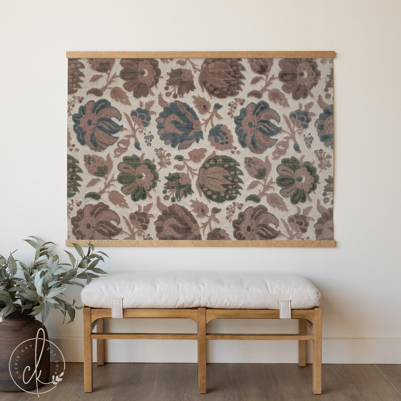 May include: Floral fabric wall hanging with a wooden frame. The fabric features a repeating pattern of large flowers in shades of brown, green, and blue on a cream background. The wooden frame is a light brown color. A wooden bench with a white cushion is below.
